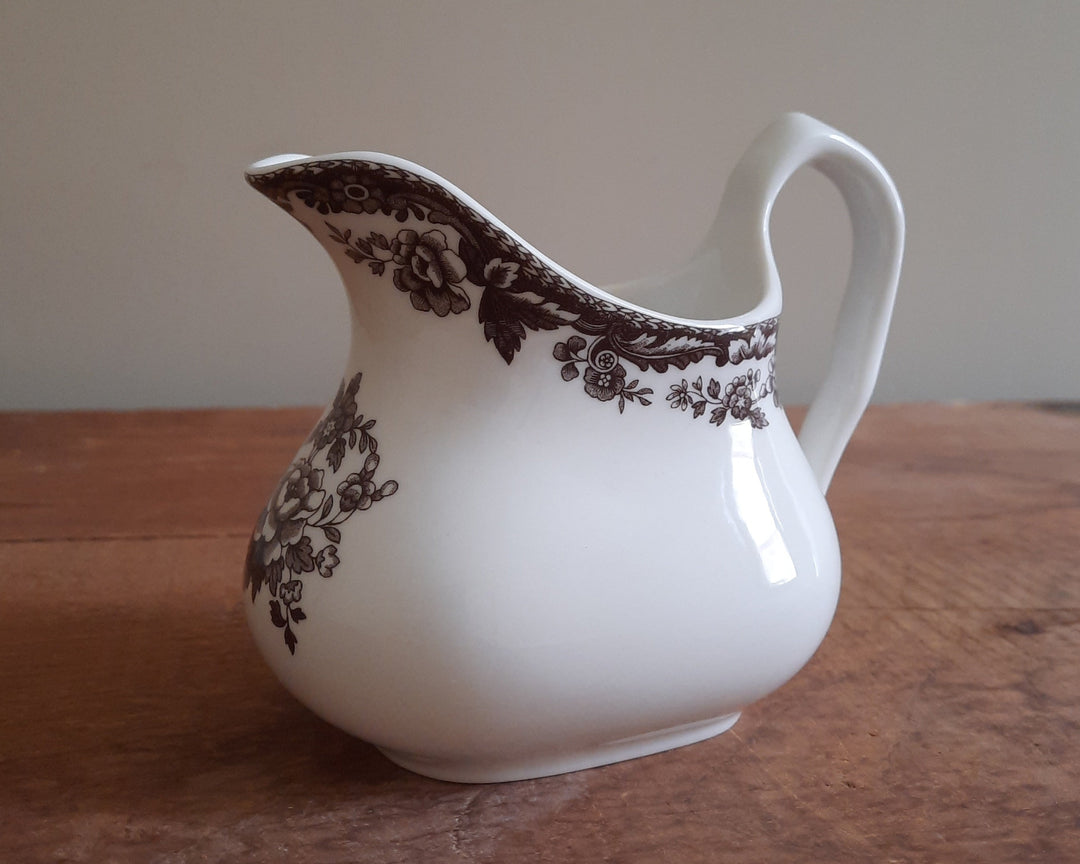 A white ceramic creamer pitcher with a dark brown floral and scroll pattern, resting on a wooden tabletop against a neutral beige background.