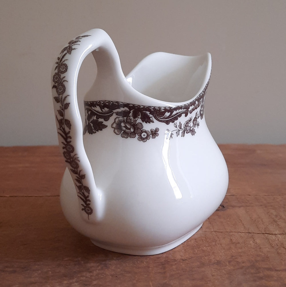 A white ceramic creamer pitcher with a dark brown floral and scroll pattern, sitting on a wooden tabletop against a neutral beige wall.