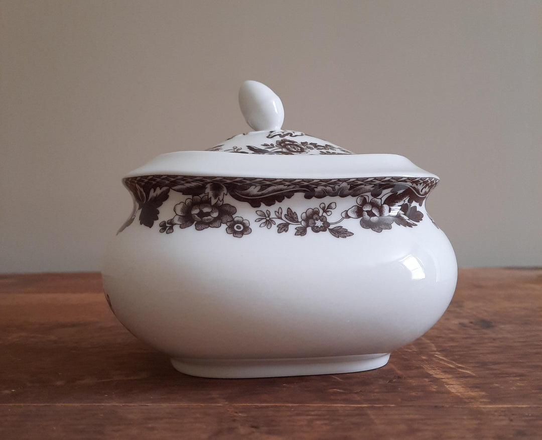A white ceramic lidded sugar bowl with a dark brown floral pattern, resting on a wooden surface against a plain beige background.