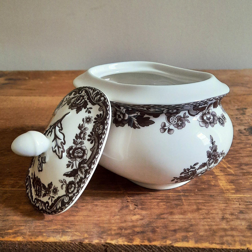 A white ceramic sugar bowl with a dark brown floral toile pattern, shown with its matching lid propped beside it on a rustic wooden table.