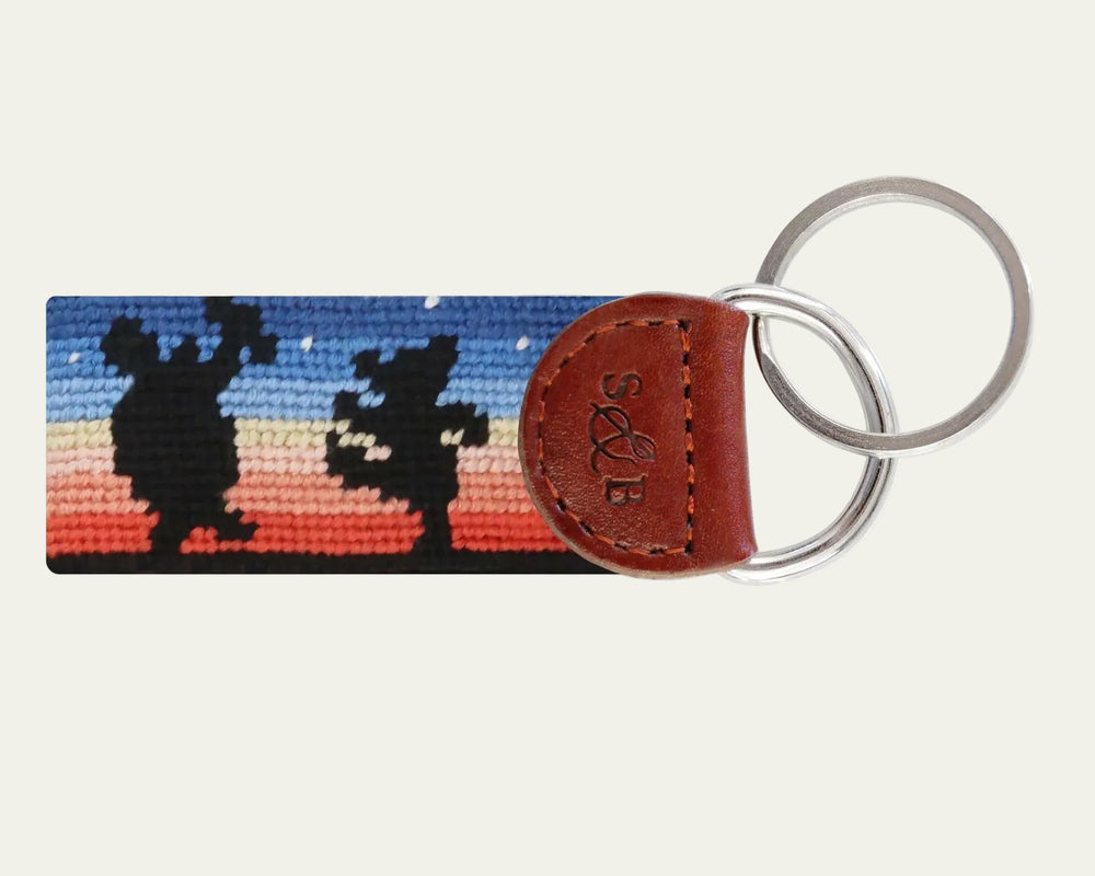 Keychain with needlepoint design of two figures against a sunset sky, attached to a brown leather tag.