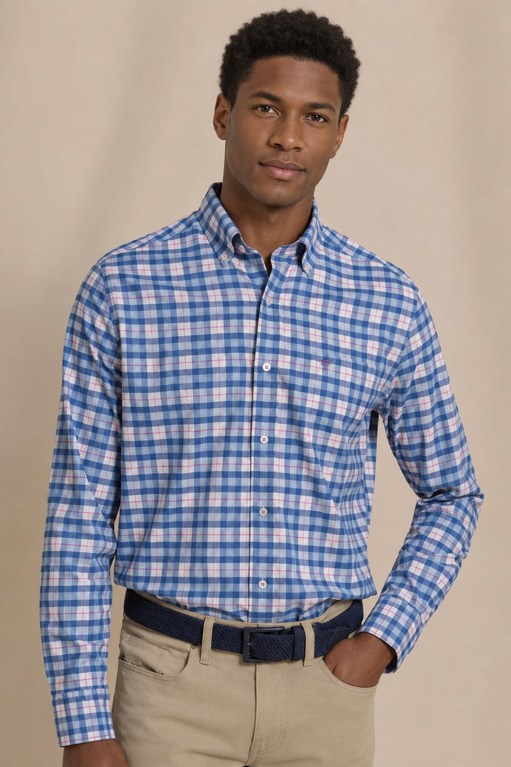 A man wearing a blue and white checkered long-sleeve button-down shirt, paired with khaki pants and a dark belt, standing against a plain beige background.