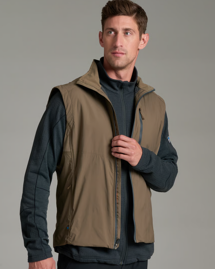 A man wearing a tan zip-front vest over a dark navy long-sleeve shirt, photographed against a plain light gray studio background.