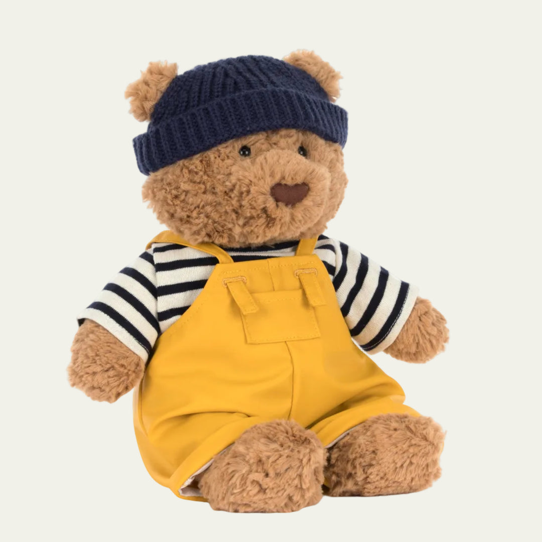 Tan plush teddy bear wearing navy knit beanie navy and white striped sweater and yellow waterproof overalls
