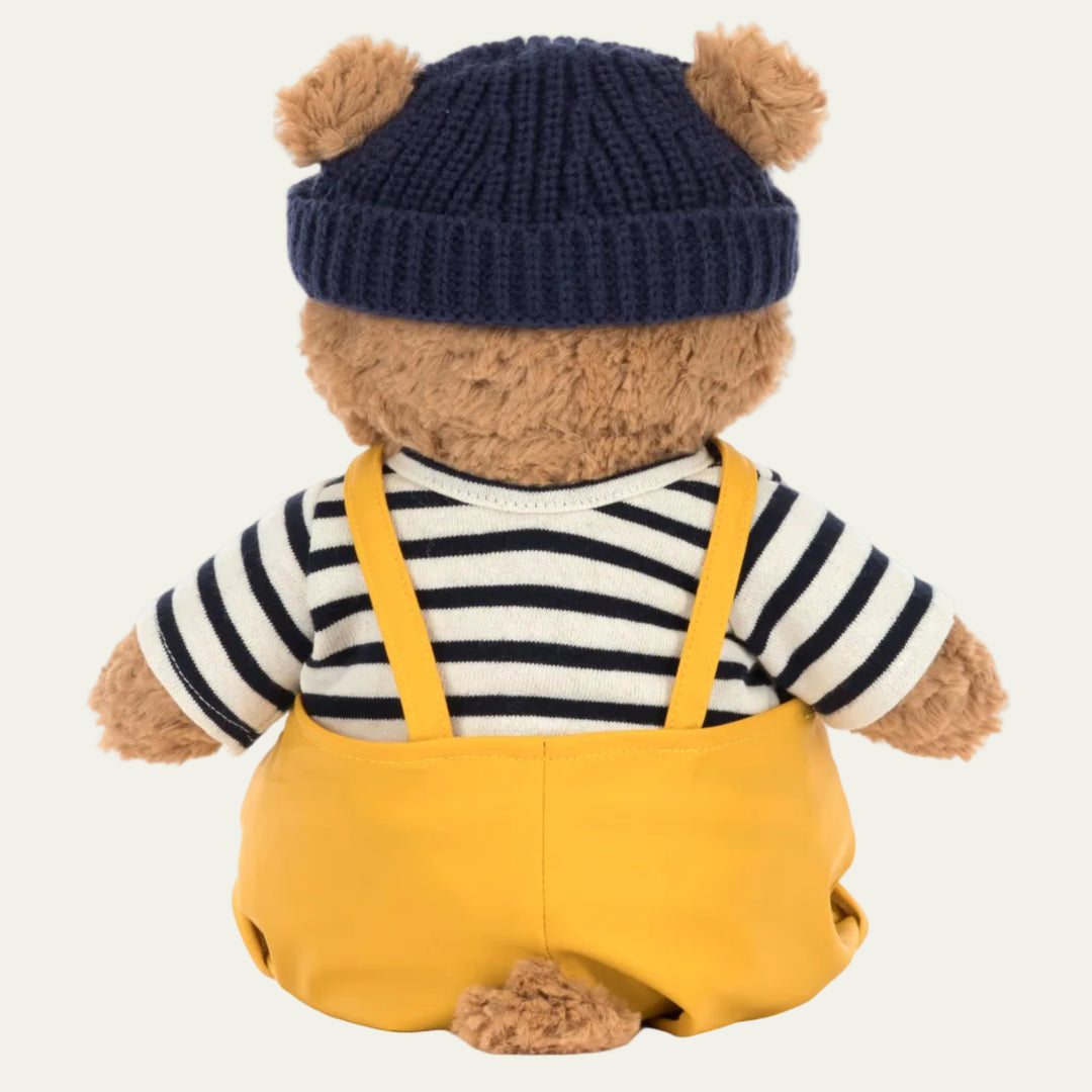 Teddy bear wearing a navy hat, striped shirt, and yellow overalls on a white background